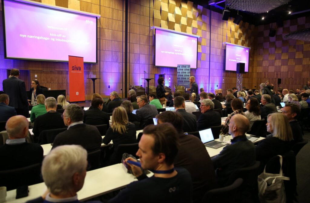 Bilde av mange mennesker i en konferansesal med tre skjermer.