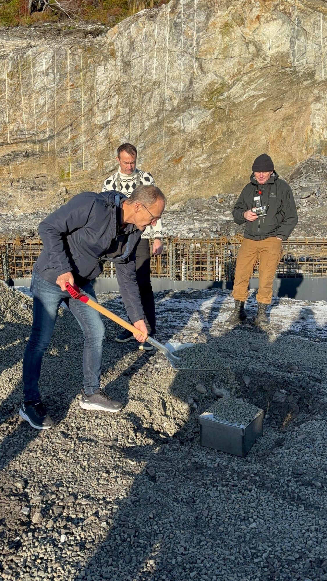 Bildet viser tre menn hvorav den ene står med en spade og skal grave ned en grunnstein. De to andre står og ser på.
