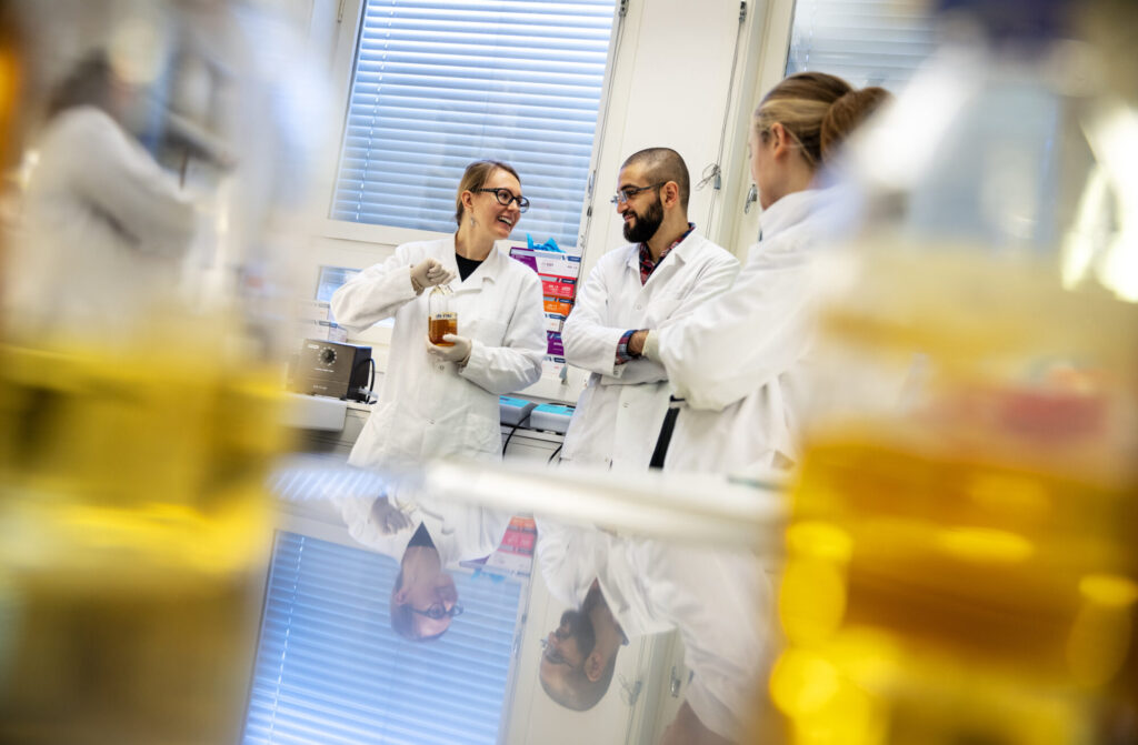 Bildet viser tre personer i hvite frakker som står i et laboratorium og prater sammen.